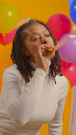 Vertical-Video-Studio-Portrait-Of-Woman-Wearing-Birthday-Headband-Celebrating-With-Balloons-And-Party-Blower